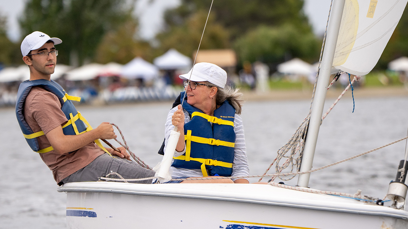 Two people in a sailboat on shoreline lake.